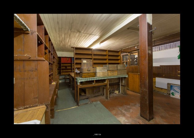 A picture of inside the shop. It has empty, dusty wooden shelves. The shop is unfurnished and an old lino floor.