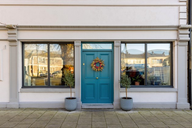 Big blue front door with an authum wreath on it. A double window is on either side of the door with a potted plant accompanying it.